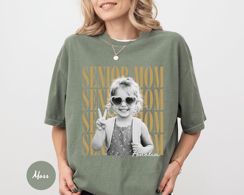 May include: A moss green short-sleeve t-shirt featuring a black and white photograph of a child wearing heart-shaped sunglasses and making a peace sign. The text SENIOR MOM is repeated in gold behind the photo, with the name Amelia in white script.