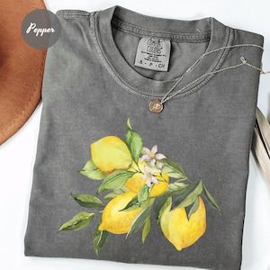 May include: A gray Comfort Colors t-shirt featuring a watercolor illustration of lemons, leaves, and small white flowers. The shirt has a crew neck and short sleeves. A silver necklace with a small pendant is draped on the shirt.
