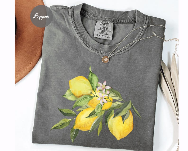May include: A gray Comfort Colors t-shirt featuring a watercolor illustration of lemons, leaves, and small white flowers. The shirt has a crew neck and short sleeves. A silver necklace with a small pendant is draped on the shirt.