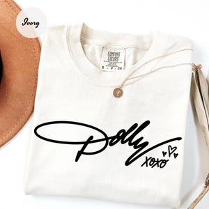 May include: An ivory short-sleeve t-shirt with a black graphic print on the front. The text reads Dolly in a cursive script, followed by XOXO and three small heart outlines. The shirt features a Comfort Colors brand label at the inner neckline.