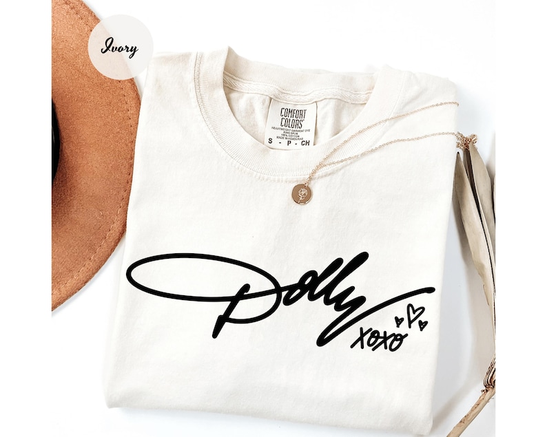 May include: An ivory short-sleeve t-shirt with a black graphic print on the front. The text reads Dolly in a cursive script, followed by XOXO and three small heart outlines. The shirt features a Comfort Colors brand label at the inner neckline.