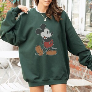 May include: Dark green sweatshirt featuring a classic Mickey Mouse graphic. The cartoon character is depicted in black, red, and tan colors. The sweatshirt has a crew neck and long sleeves, perfect for casual wear.