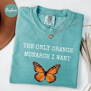 Pode incluir: Camiseta verde-&aacute;gua com o texto "THE ONLY ORANGE MONARCH I WANT" e um gr&aacute;fico de borboleta monarca. Um colar de ouro est&aacute; drapeado sobre a camiseta. A camiseta est&aacute; dobrada sobre uma superf&iacute;cie branca.