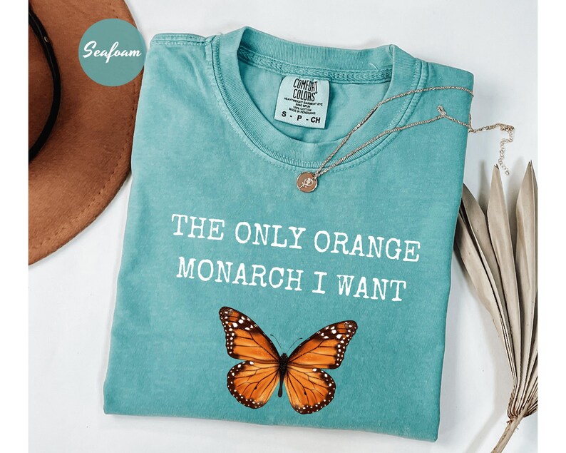 Pode incluir: Camiseta verde-&aacute;gua com o texto "THE ONLY ORANGE MONARCH I WANT" e um gr&aacute;fico de borboleta monarca. Um colar de ouro est&aacute; drapeado sobre a camiseta. A camiseta est&aacute; dobrada sobre uma superf&iacute;cie branca.