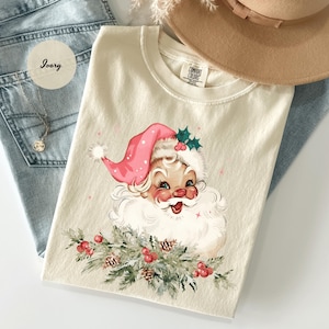 May include: Ivory-colored t-shirt featuring a vintage Santa Claus graphic. Santa wears a pink hat and has a white beard. The graphic includes holly and berries. The shirt is laid on top of blue jeans and near a tan hat.