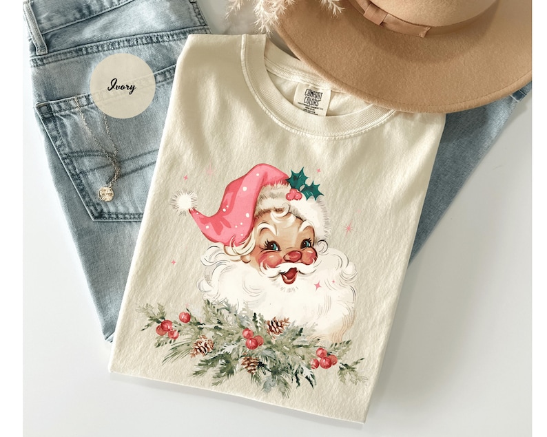 May include: Ivory-colored t-shirt featuring a vintage Santa Claus graphic. Santa wears a pink hat and has a white beard. The graphic includes holly and berries. The shirt is laid on top of blue jeans and near a tan hat.