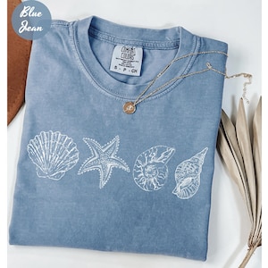 May include: A blue jean Comfort Colors t-shirt with a white seashell design. The design includes a seashell, starfish, and two other shell illustrations. A gold necklace with a pendant is draped across the shirt.