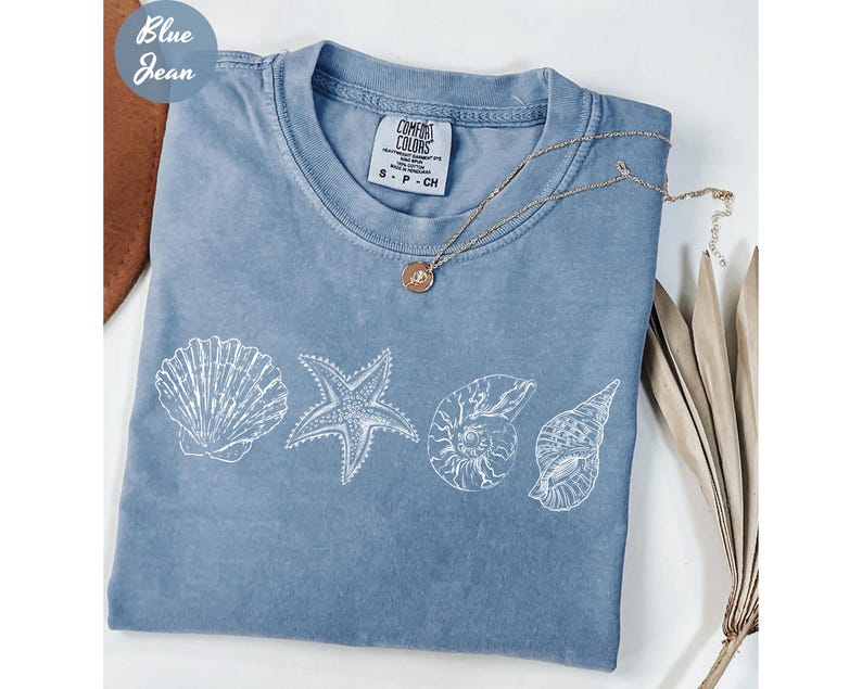 May include: A blue jean Comfort Colors t-shirt with a white seashell design. The design includes a seashell, starfish, and two other shell illustrations. A gold necklace with a pendant is draped across the shirt.