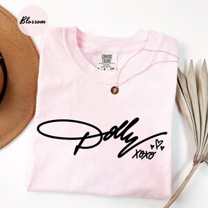 May include: A light pink short-sleeve t-shirt featuring the black script text Dolly and the word xoxo with three small heart outlines. The shirt has a crew neckline and a Comfort Colors brand label at the inner collar, size S-CH.