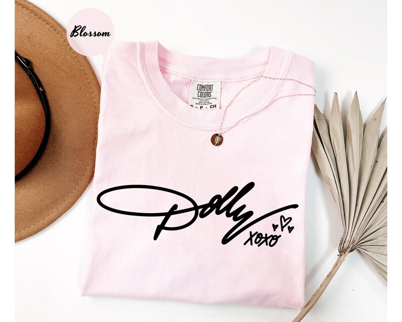 May include: A light pink short-sleeve t-shirt featuring the black script text Dolly and the word xoxo with three small heart outlines. The shirt has a crew neckline and a Comfort Colors brand label at the inner collar, size S-CH.