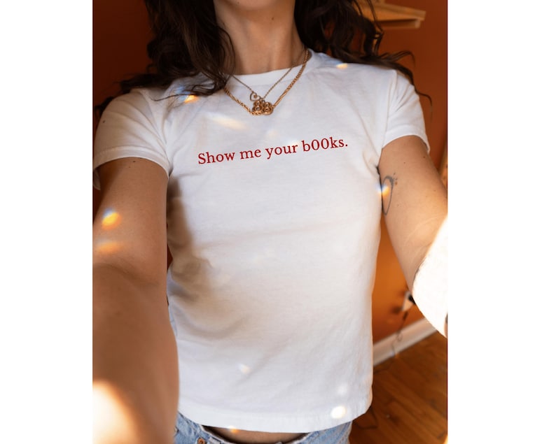 May include: White t-shirt with the red text "Show me your books." The shirt has short sleeves and a crew neck. A gold necklace is visible. The model is wearing blue jeans.