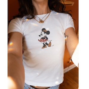 Pu&ograve; includere: T-shirt bianco con grafica di Minnie Mouse. Il design mostra Minnie con una gonna a pois rossi, scarpe marroni e un fiocco nero. La maglietta ha maniche corte e girocollo.