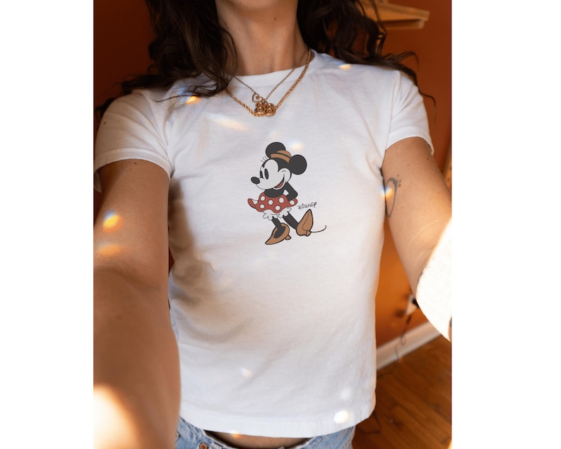 Pu&ograve; includere: T-shirt bianco con grafica di Minnie Mouse. Il design mostra Minnie con una gonna a pois rossi, scarpe marroni e un fiocco nero. La maglietta ha maniche corte e girocollo.