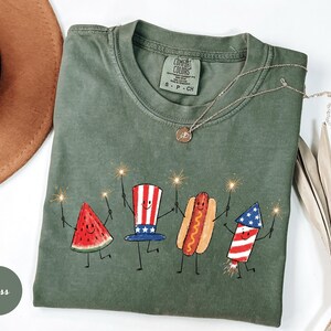 K&ouml;nnte beinhalten: Moosgr&uuml;nes Comfort Colors T-Shirt mit einer verspielten Illustration einer Wassermelonenscheibe, einem Hotdog und einem Feuerwerk, die jeweils Wunderkerzen halten. Das Shirt hat einen Rundhalsausschnitt und kurze &Auml;rmel. Eine goldene Halskette mit Anh&auml;nger befindet sich auf dem Shirt.