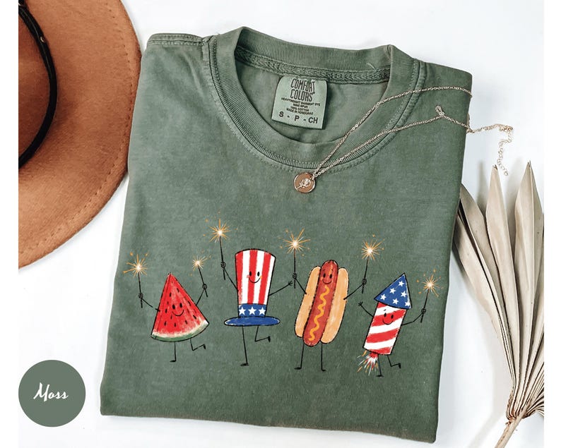 K&ouml;nnte beinhalten: Moosgr&uuml;nes Comfort Colors T-Shirt mit einer verspielten Illustration einer Wassermelonenscheibe, einem Hotdog und einem Feuerwerk, die jeweils Wunderkerzen halten. Das Shirt hat einen Rundhalsausschnitt und kurze &Auml;rmel. Eine goldene Halskette mit Anh&auml;nger befindet sich auf dem Shirt.