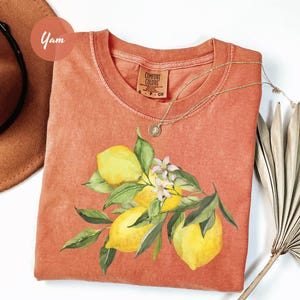 May include: A coral-colored t-shirt featuring a watercolor illustration of lemons and leaves. The design includes yellow lemons, green leaves, and small white flowers. A gold necklace is draped over the shirt.