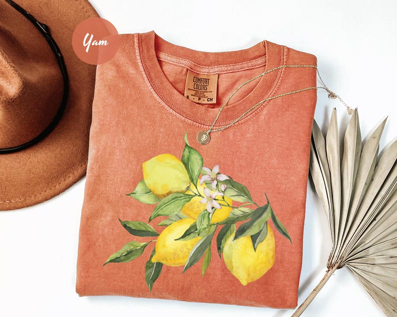 May include: A coral-colored t-shirt featuring a watercolor illustration of lemons and leaves. The design includes yellow lemons, green leaves, and small white flowers. A gold necklace is draped over the shirt.