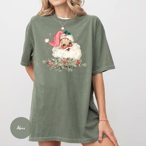 May include: An olive green t-shirt featuring a vintage Santa Claus graphic. The Santa illustration has a pink hat, white beard, and is surrounded by festive greenery and red berries. The shirt is a loose fit.