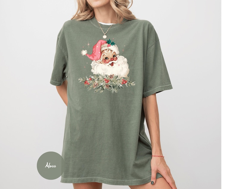 May include: An olive green t-shirt featuring a vintage Santa Claus graphic. The Santa illustration has a pink hat, white beard, and is surrounded by festive greenery and red berries. The shirt is a loose fit.