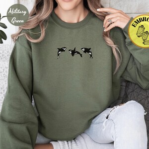 May include: Military green sweatshirt with three embroidered black and white orcas. The sweatshirt has long sleeves and a crew neck. The text "Embroidery" is on a yellow circle. The words "Military Green" are on a white circle.