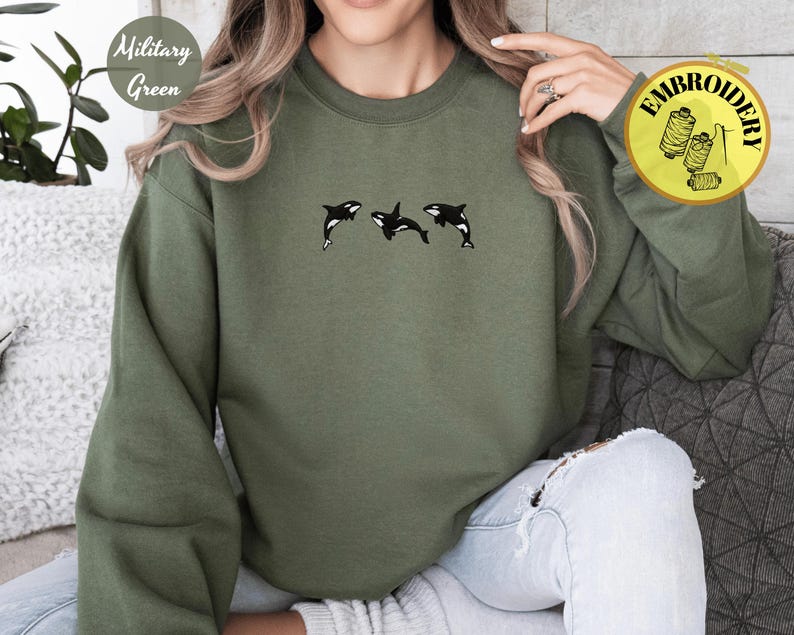 May include: Military green sweatshirt with three embroidered black and white orcas. The sweatshirt has long sleeves and a crew neck. The text "Embroidery" is on a yellow circle. The words "Military Green" are on a white circle.