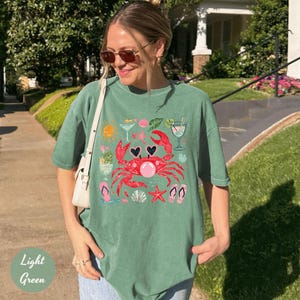 May include: Light green t-shirt with a graphic of a red crab wearing sunglasses, surrounded by beach-themed icons. The shirt also features the text "Light Green" in a light green font.