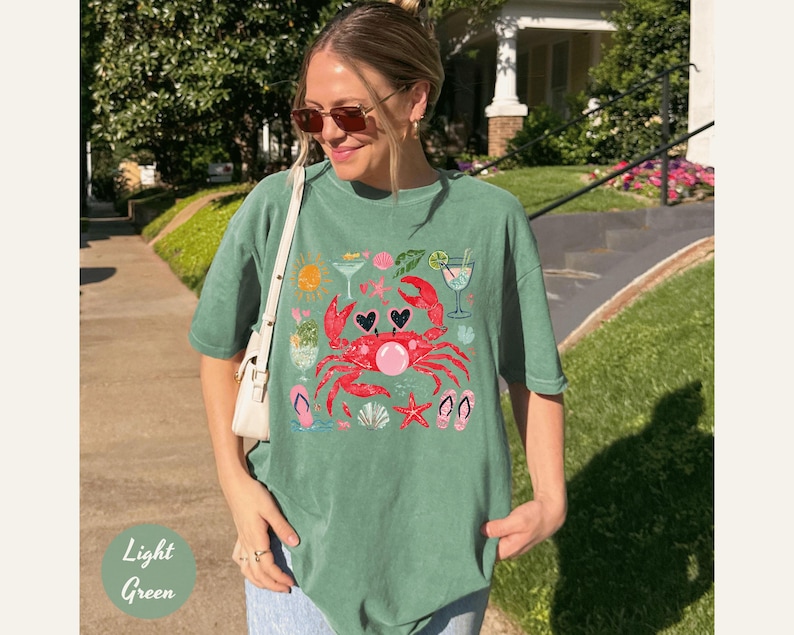 May include: Light green t-shirt with a graphic of a red crab wearing sunglasses, surrounded by beach-themed icons. The shirt also features the text "Light Green" in a light green font.