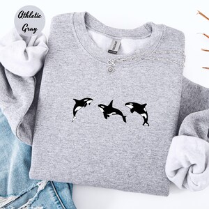 May include: Athletic gray sweatshirt featuring three black and white orca whale graphics. The sweatshirt has a crew neck and long sleeves. The text "Athletic Gray" is in a circle in the upper left corner.