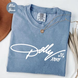 May include: A blue jean colored t-shirt with a crew neckline and a white graphic print on the front. The print features the word Dolly in a cursive script font, followed by the text xoxo and three small heart outlines. The shirt has a tag.