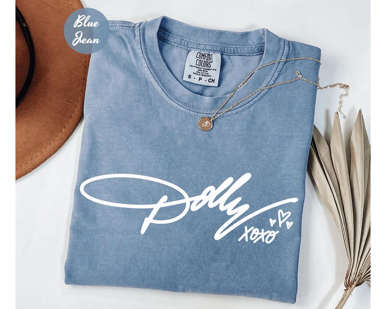 May include: A blue jean colored t-shirt with a crew neckline and a white graphic print on the front. The print features the word Dolly in a cursive script font, followed by the text xoxo and three small heart outlines. The shirt has a tag.
