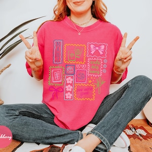 May include: A bright pink t-shirt with a collection of colorful stamp-like designs. The stamps feature various graphics, including a bow, flowers, and the words "MAUI" and "one more game". The person is wearing blue jeans.