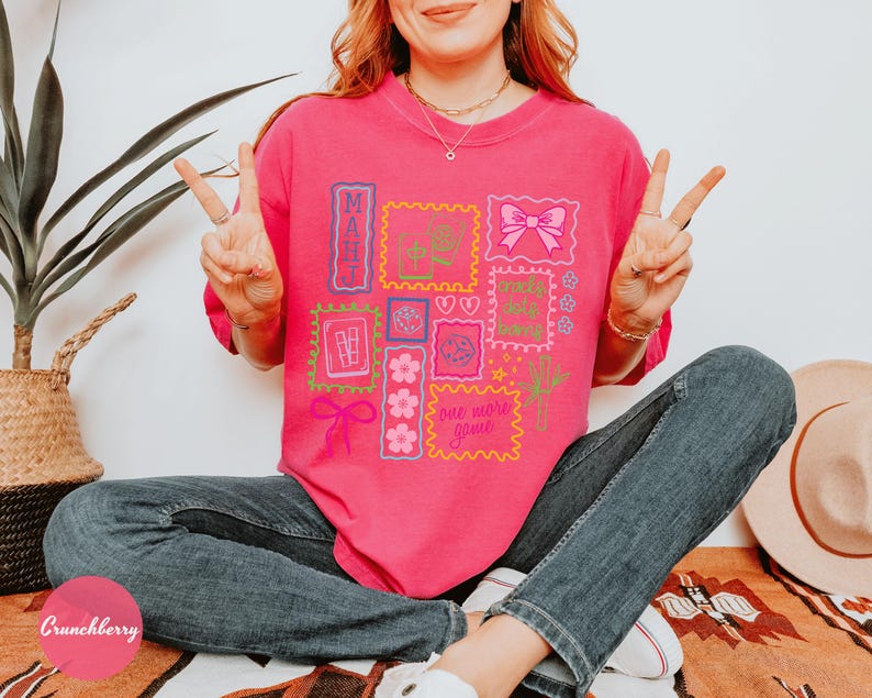 May include: A bright pink t-shirt with a collection of colorful stamp-like designs. The stamps feature various graphics, including a bow, flowers, and the words "MAUI" and "one more game". The person is wearing blue jeans.