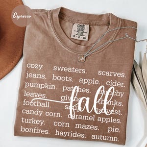 May include: An espresso-colored t-shirt with a crew neckline and a Comfort Colors label. The shirt features white text listing fall-themed words including sweaters, scarves, jeans, boots, apple cider, pumpkin, patches, leaves, football, and autumn.