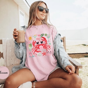 May include: Pink t-shirt with a crab graphic, heart-shaped eyes, and a bubble. The design includes a sun, cocktails, and starfish. The shirt is worn with sunglasses and a denim jacket.