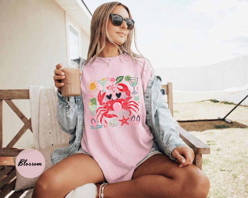 May include: Pink t-shirt with a crab graphic, heart-shaped eyes, and a bubble. The design includes a sun, cocktails, and starfish. The shirt is worn with sunglasses and a denim jacket.