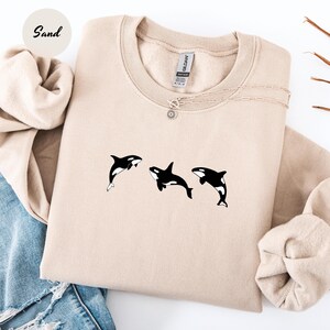 May include: A sand-colored sweatshirt featuring a crew neck and long sleeves. The sweatshirt has a graphic of three black and white orcas. A delicate silver necklace with a sun pendant is visible.