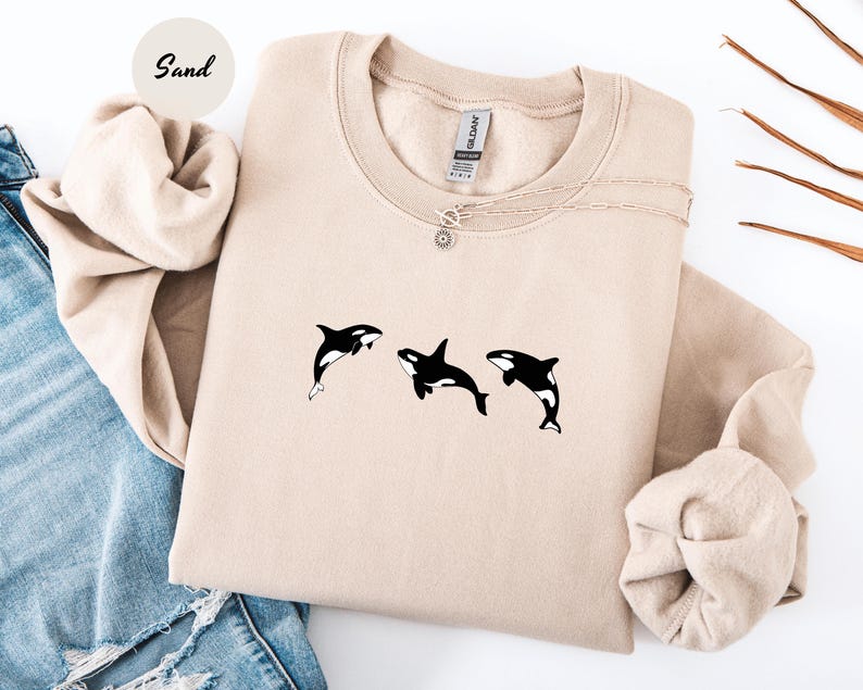 May include: A sand-colored sweatshirt featuring a crew neck and long sleeves. The sweatshirt has a graphic of three black and white orcas. A delicate silver necklace with a sun pendant is visible.