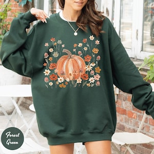 May include: A forest green long-sleeve sweatshirt featuring a central graphic of a large orange pumpkin surrounded by a variety of small, colorful wildflowers in shades of red, orange, yellow, and white. The garment has a crew neckline.