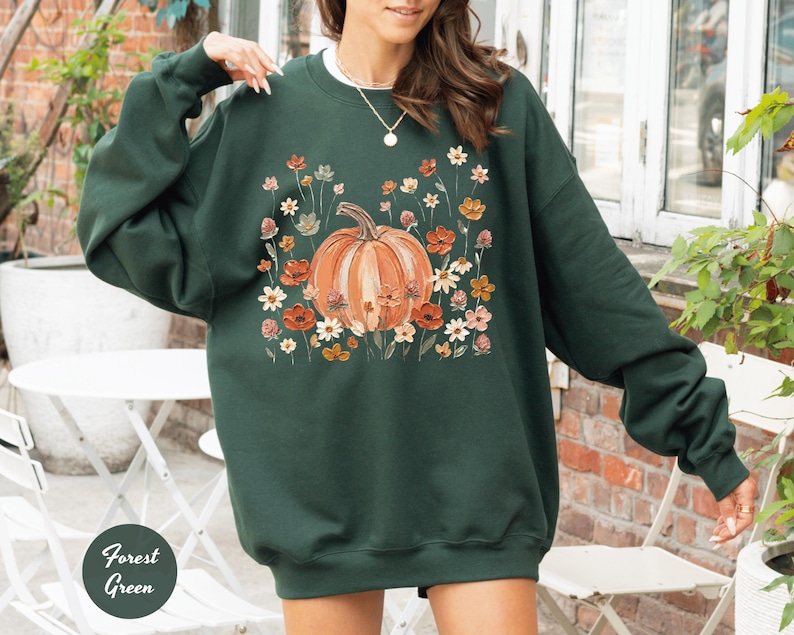 May include: A forest green long-sleeve sweatshirt featuring a central graphic of a large orange pumpkin surrounded by a variety of small, colorful wildflowers in shades of red, orange, yellow, and white. The garment has a crew neckline.