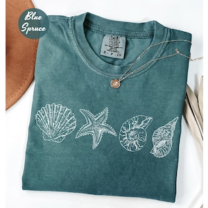 May include: A blue spruce t-shirt featuring white seashell illustrations. The shirt has a crew neck and short sleeves. A gold necklace with a pendant rests on the shirt. The shirt is labeled "Comfort Colors".