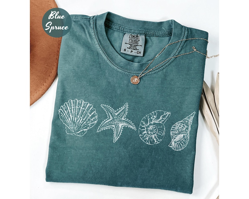 May include: A blue spruce t-shirt featuring white seashell illustrations. The shirt has a crew neck and short sleeves. A gold necklace with a pendant rests on the shirt. The shirt is labeled "Comfort Colors".
