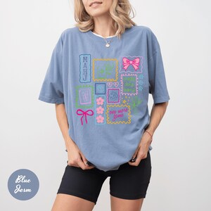 May include: A light blue t-shirt with a colorful graphic design featuring various framed elements, including a pink bow, flowers, and the text "one more game". The shirt is a comfortable, oversized fit.