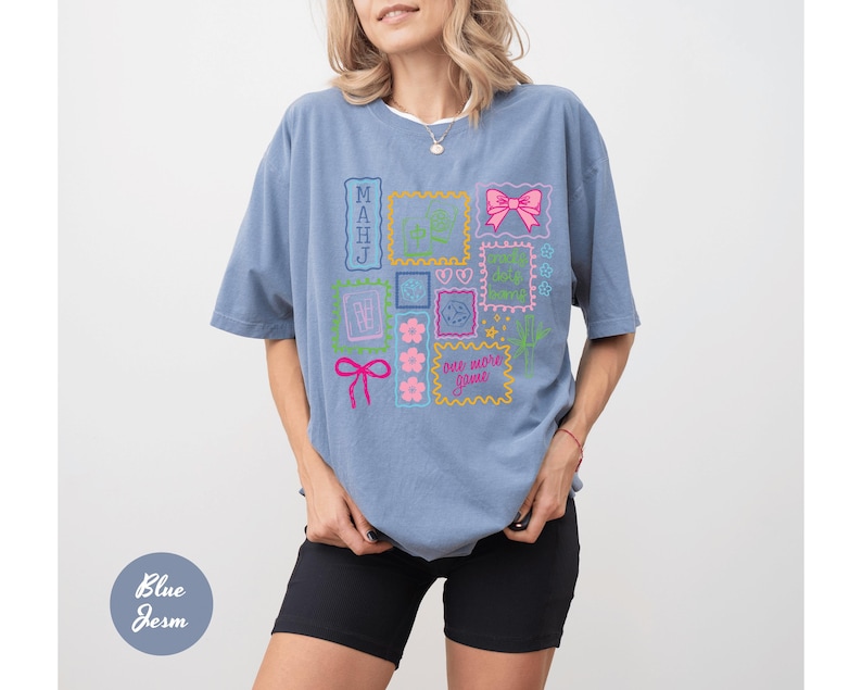 May include: A light blue t-shirt with a colorful graphic design featuring various framed elements, including a pink bow, flowers, and the text "one more game". The shirt is a comfortable, oversized fit.
