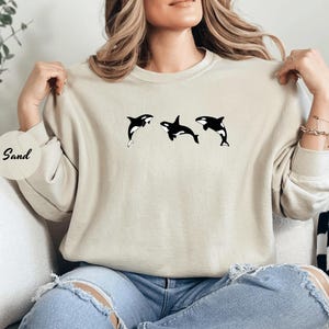 May include: A sand-colored sweatshirt featuring three black and white orca whale graphics. The orcas are depicted in various poses, with one leaping out of the water. The word "Sand" is printed on a round sticker.