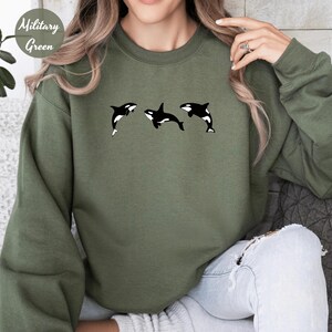 May include: A military green sweatshirt featuring three black and white orca whale graphics. The sweatshirt has long sleeves and a crew neck. The words "Military Green" are printed in a circle in the upper left corner.