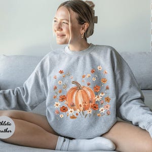 May include: An athletic heather gray crewneck sweatshirt featuring a central graphic print of a large orange pumpkin surrounded by a variety of small orange, white, and brown flowers and foliage. The sweatshirt has long sleeves and a ribbed neckline.