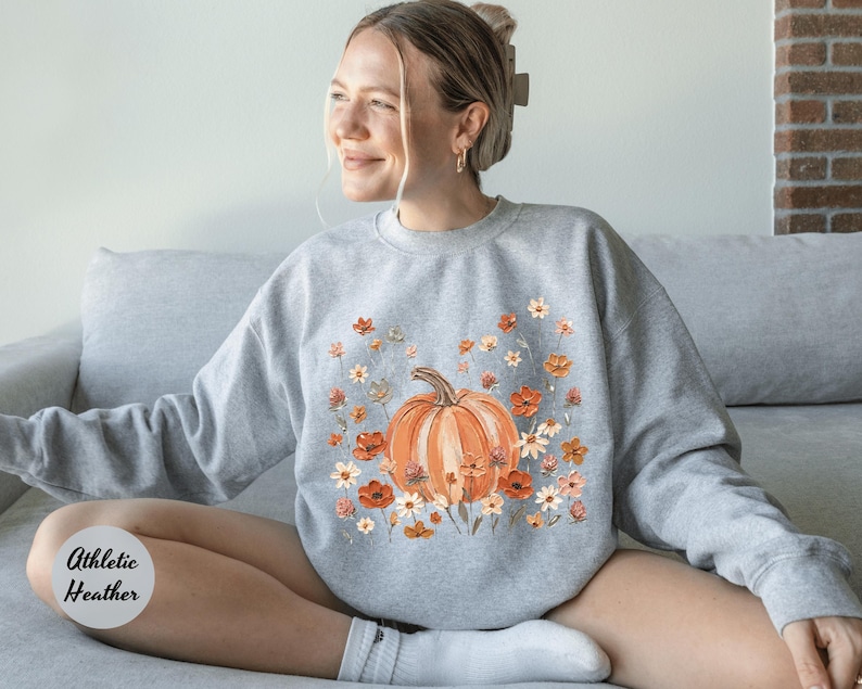 May include: An athletic heather gray crewneck sweatshirt featuring a central graphic print of a large orange pumpkin surrounded by a variety of small orange, white, and brown flowers and foliage. The sweatshirt has long sleeves and a ribbed neckline.