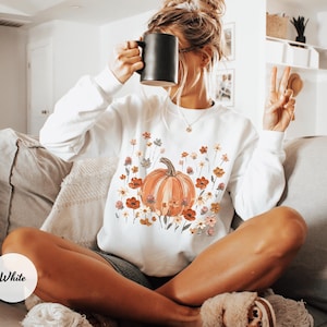 May include: A white long-sleeve sweatshirt featuring a central graphic of an orange pumpkin surrounded by a variety of colorful wildflowers in shades of red, orange, and muted green. The sweatshirt has a crew neckline and ribbed cuffs.