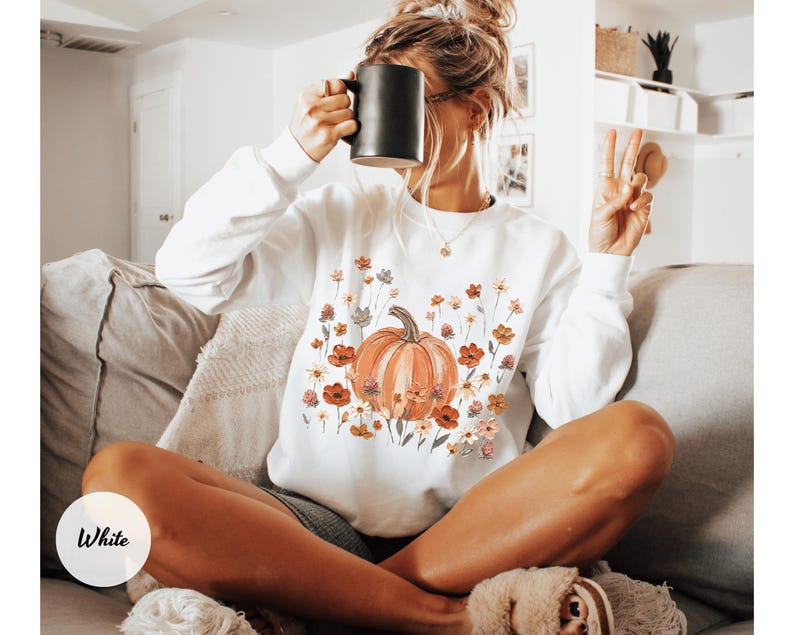 May include: A white long-sleeve sweatshirt featuring a central graphic of an orange pumpkin surrounded by a variety of colorful wildflowers in shades of red, orange, and muted green. The sweatshirt has a crew neckline and ribbed cuffs.