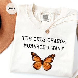Pode incluir: Camiseta cor marfim com o texto "THE ONLY ORANGE MONARCH I WANT" acima de um gr&aacute;fico detalhado de uma borboleta laranja e preta. Um colar de ouro com um pingente est&aacute; drapeado sobre a camiseta.